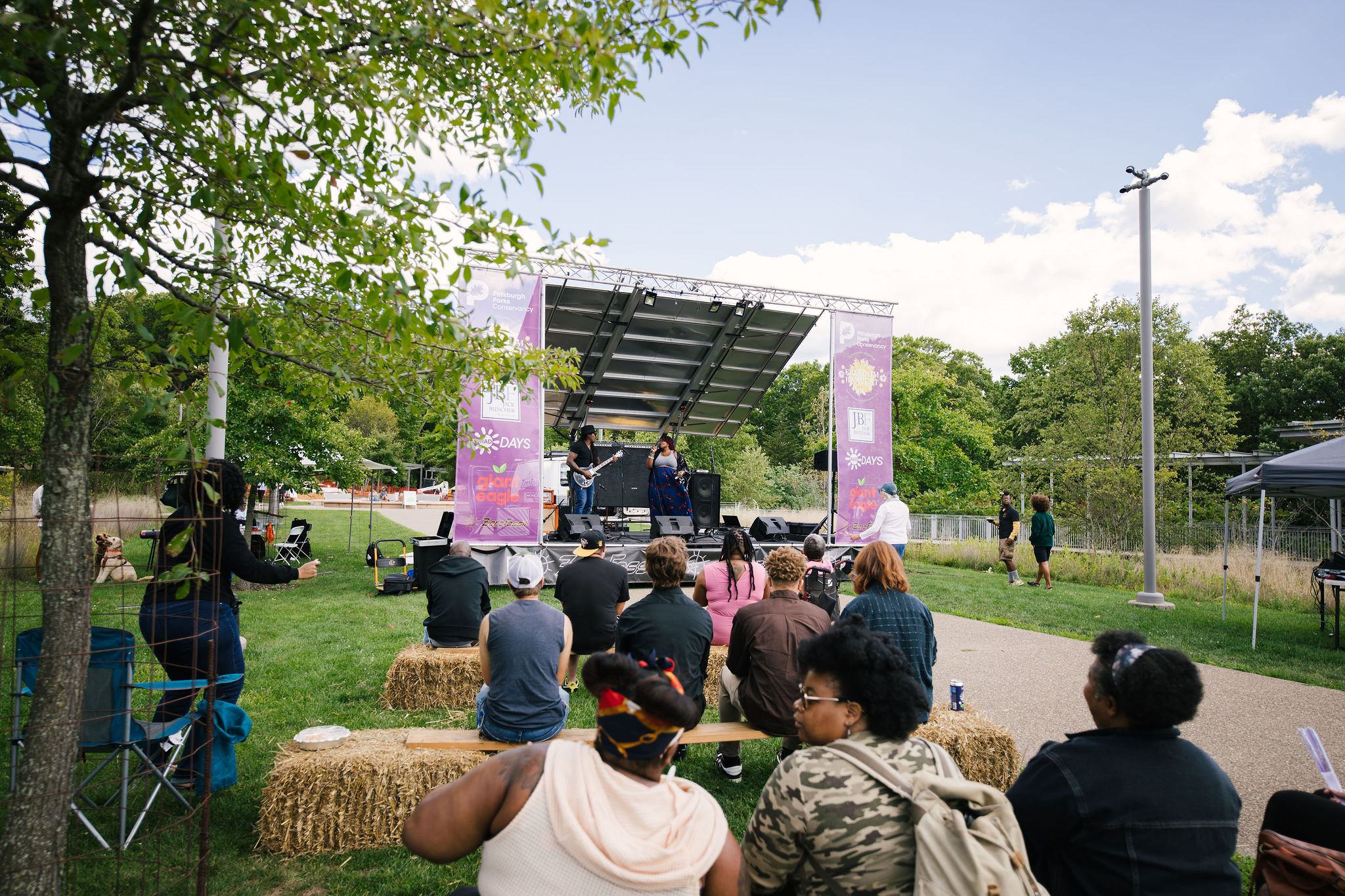 A group of people sit on hay bales and enjoy live music during Freedom Harvest Fest in Frick Park.