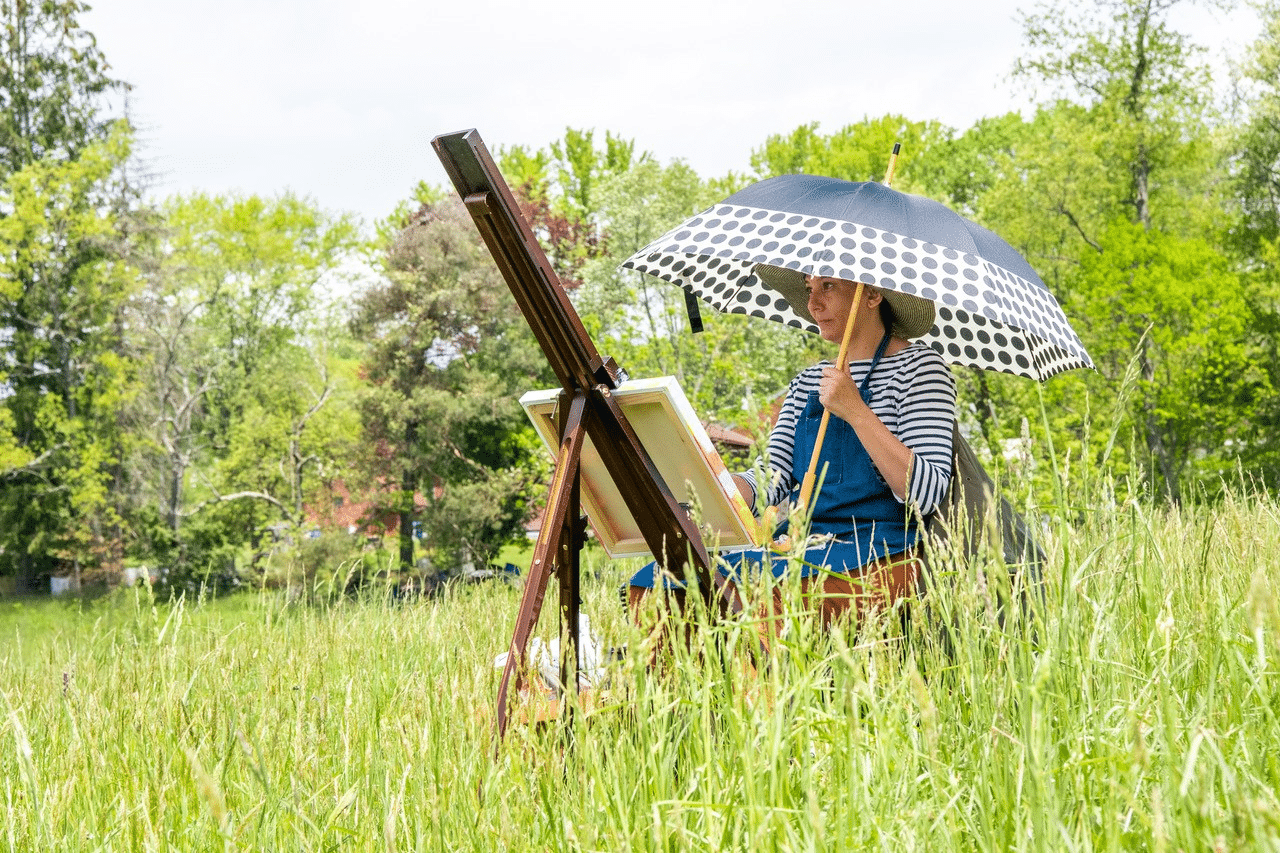 A person paints outside amongst tall grass, while holding an umbrella to protect themselves from the sun.