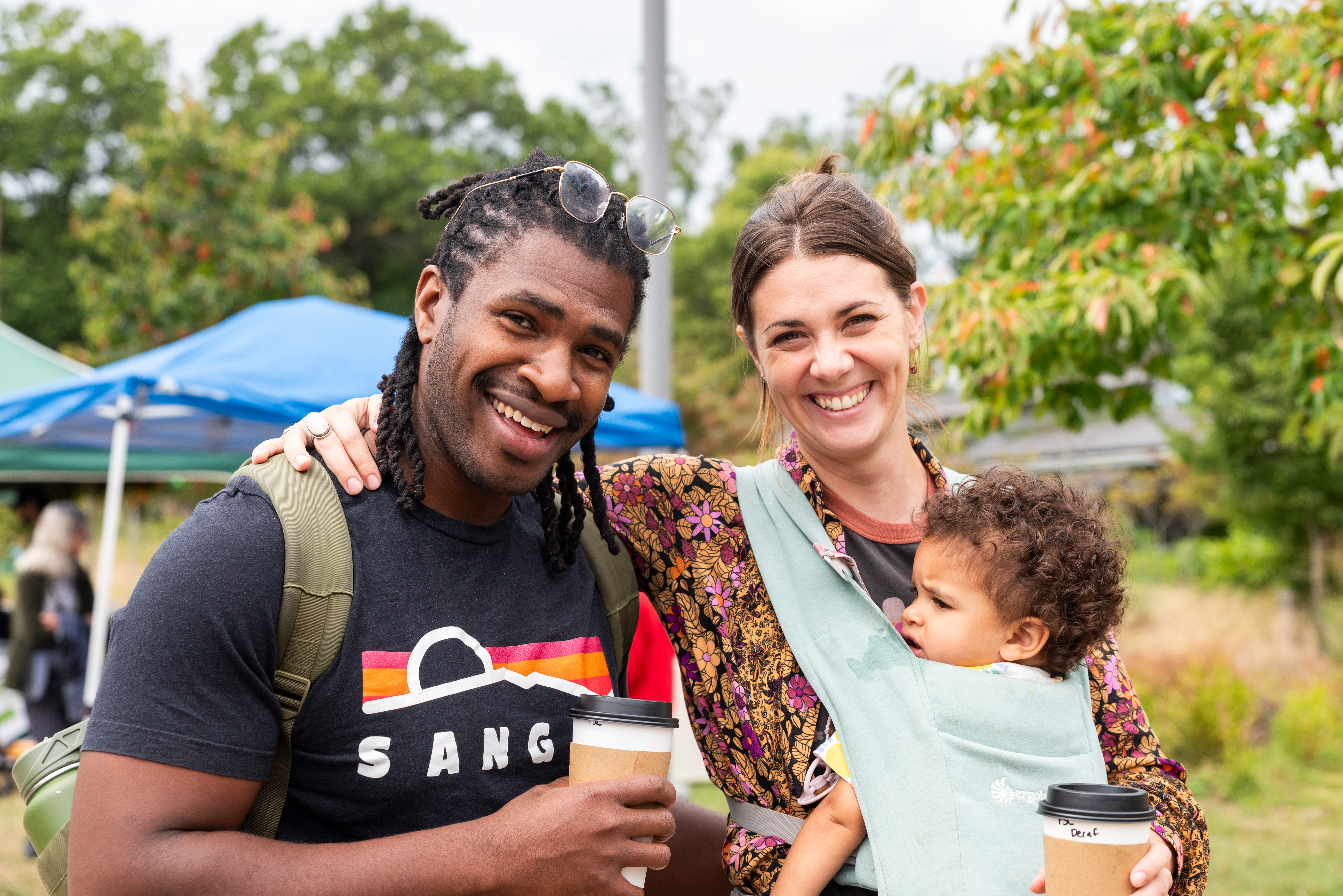 A man and a woman hold a baby while smiling in Frick Park.