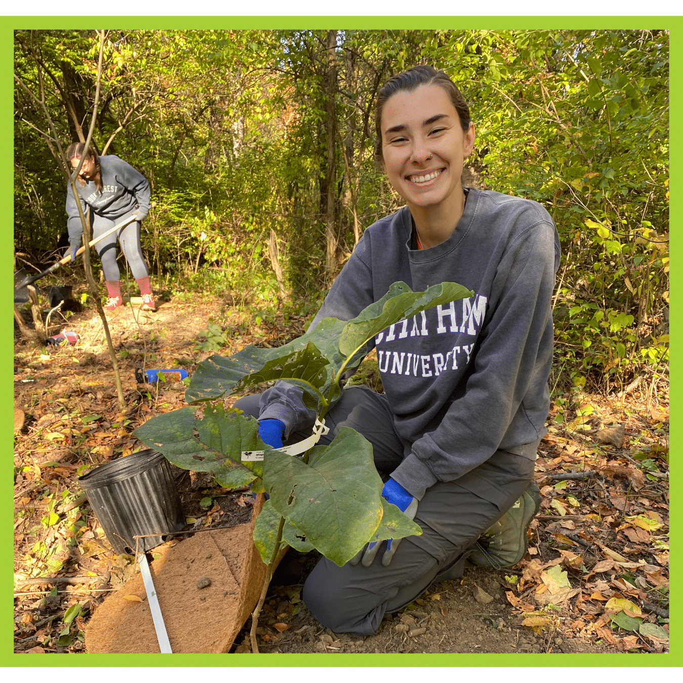 A woman kneels next to a young tree planting and smiles at the camera.
