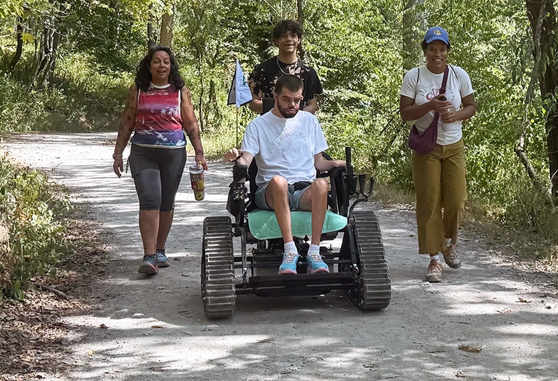 A group of park visitors explore the trails of Frick Park, with one user riding in a Trackchair.