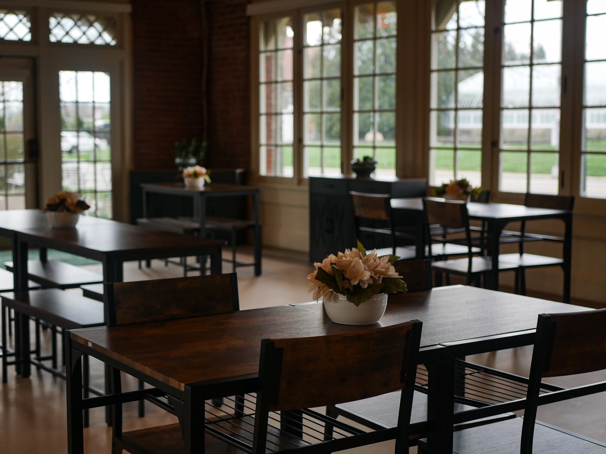Inside of a quaint building with big windows, there are rectangular tables with floral arrangements.